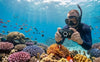 A snorkeler uses a rugged, waterproof Ricoh WG-8 camera to capture a close-up of a clownfish in an anemone on a vibrant coral reef, under natural sunlight.