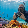 A snorkeler uses a rugged, waterproof Ricoh WG-8 camera to capture a close-up of a clownfish in an anemone on a vibrant coral reef, under natural sunlight.