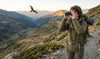 A hiker standing on a mountain trail uses a pair of black compact binoculars to observe a distant bird of prey soaring over the valley.